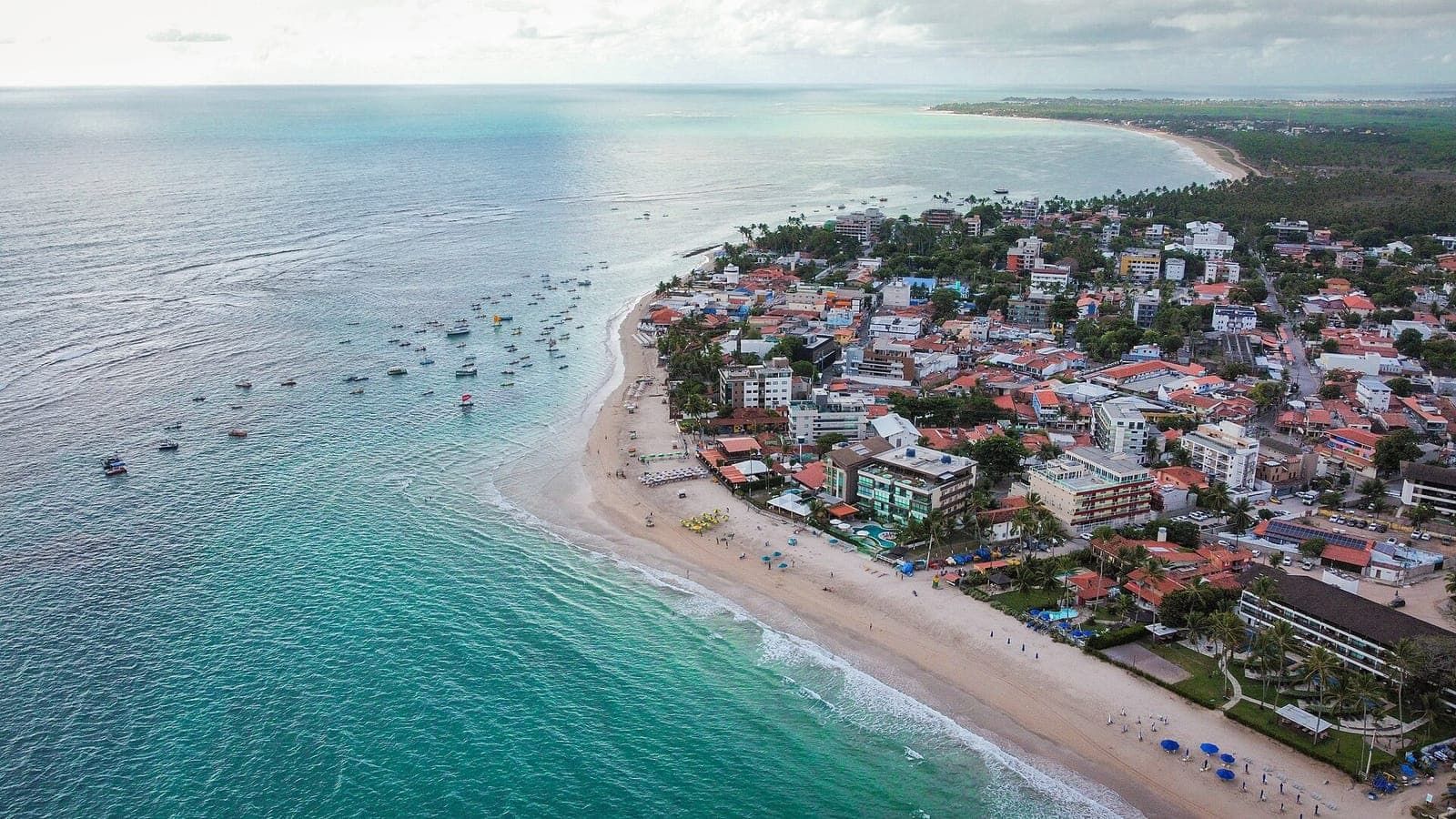 Vista aérea da praia central de Porto de Galinhas, com barcos no mar verde-água e a vila na orla