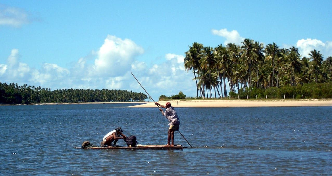 Pescadores numa jangada sobre o mar calmo de Tamandaré, com coqueiral e faixa de areia ao fundo